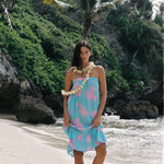A woman stands on a sandy beach wearing a light blue halter dress with pink tropical floral patterns, a white flower lei necklace, and a matching flower armband, with palm trees and rocks in the background.