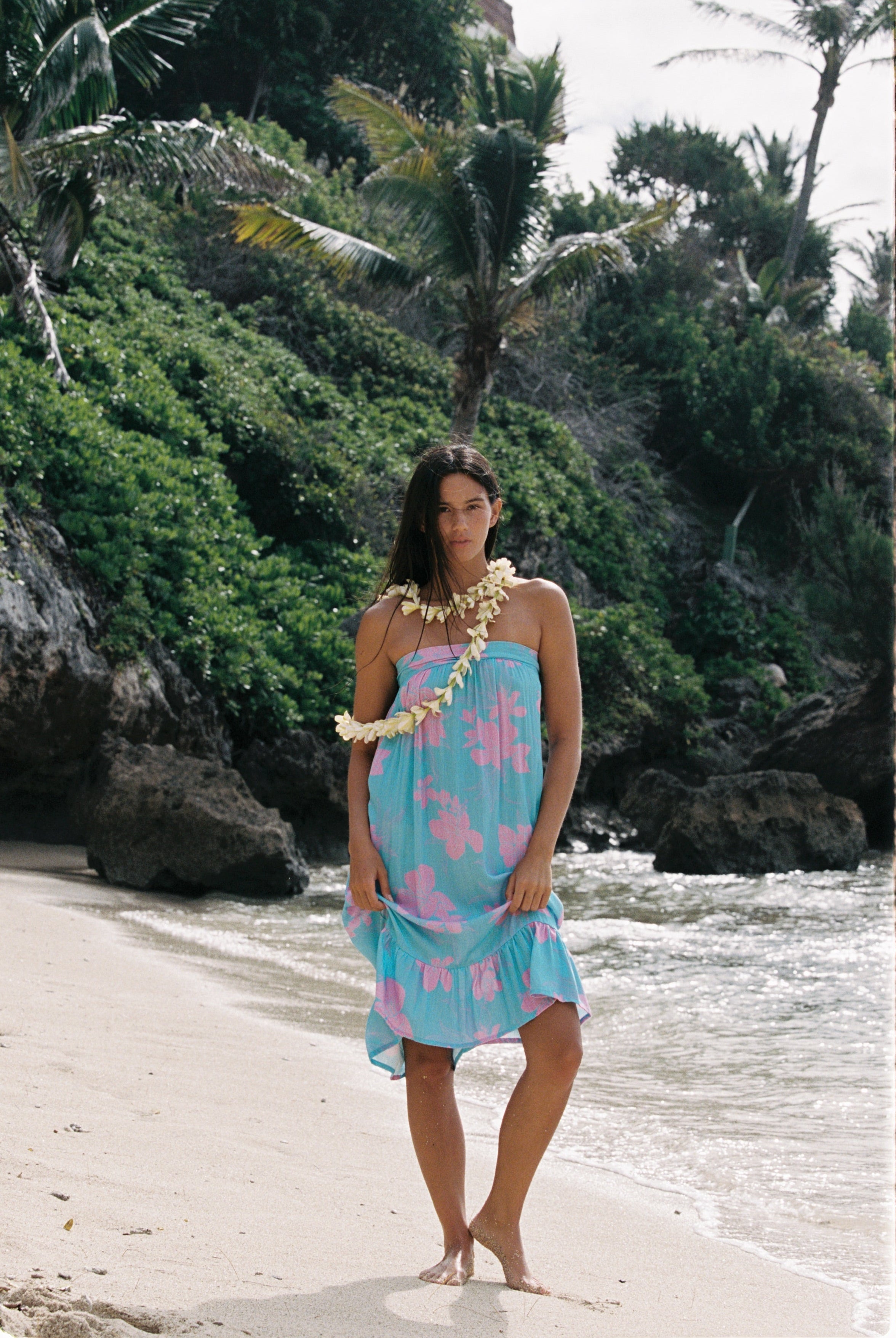 A woman stands on a sandy beach wearing a light blue halter dress with pink tropical floral patterns, a white flower lei necklace, and a matching flower armband, with palm trees and rocks in the background.