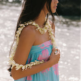 A woman on a beach wearing a strapless light blue and pink floral dress with a white flower lei necklace and matching flower arm garlands.