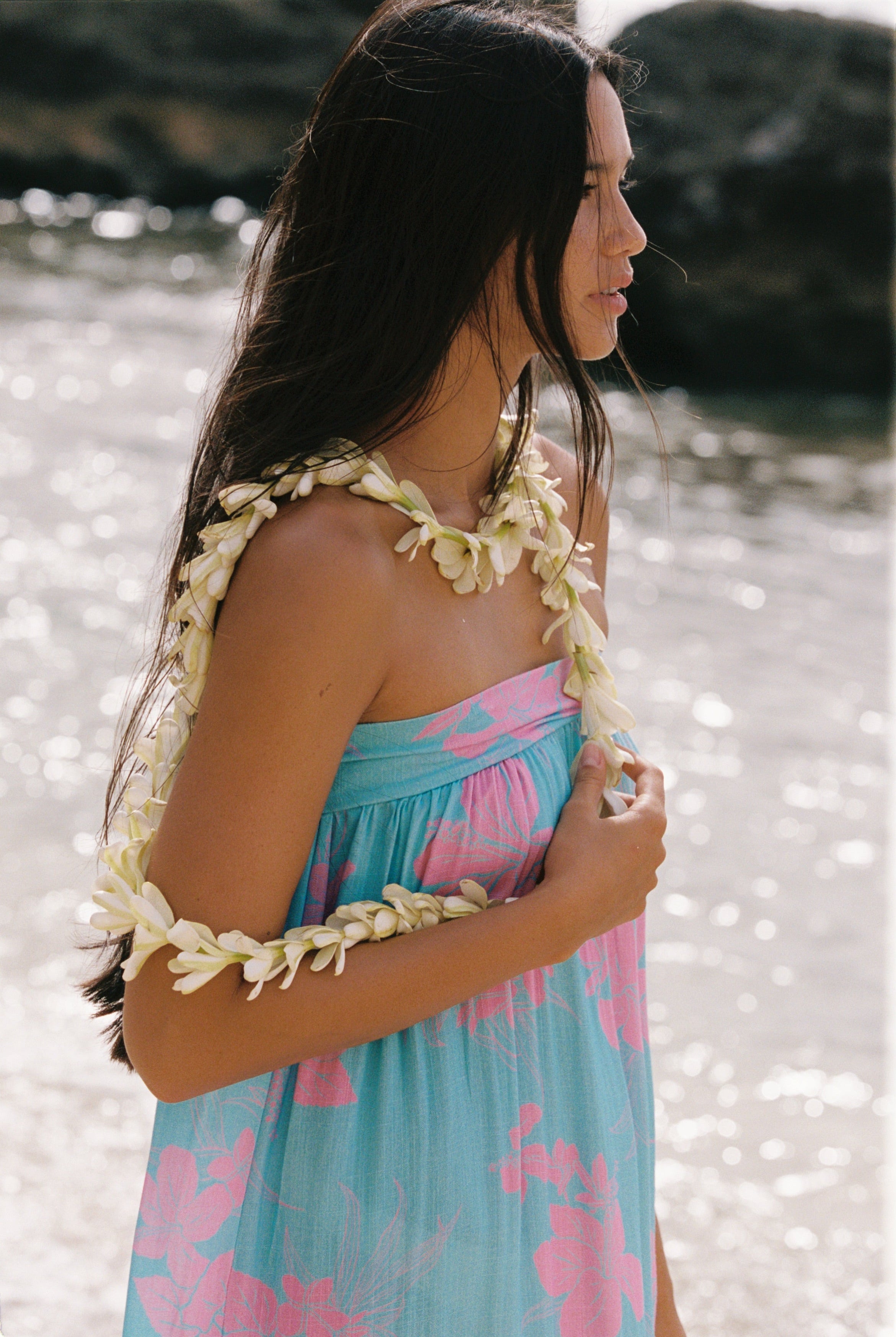 A woman on a beach wearing a strapless light blue and pink floral dress with a white flower lei necklace and matching flower arm garlands.