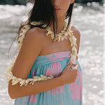 A woman wearing a light blue and pink floral strapless dress with white flower lei accessories, standing outdoors in front of a bright, sunlit water background.