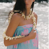 A woman wearing a light blue and pink floral strapless dress with white flower lei accessories, standing outdoors in front of a bright, sunlit water background.