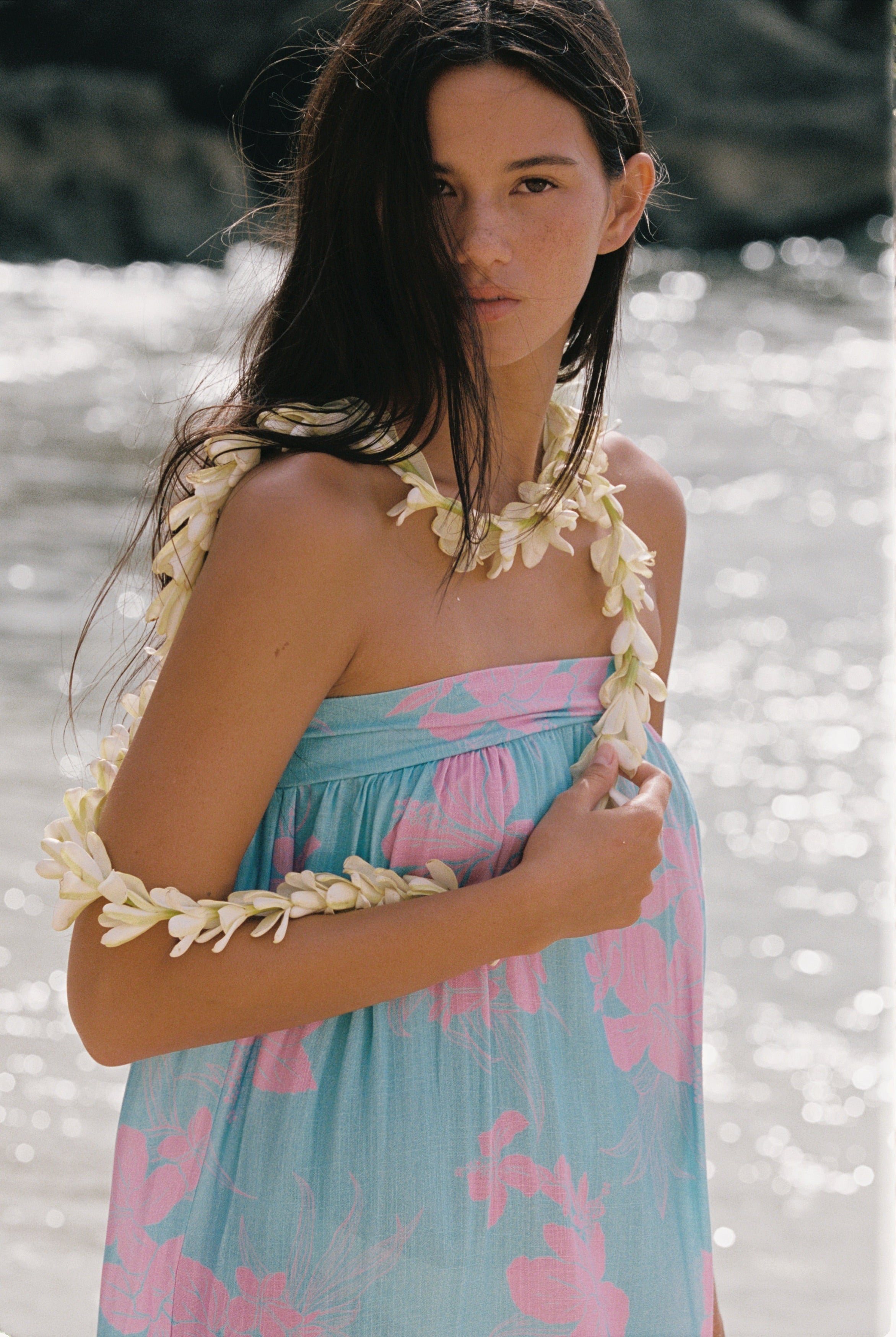 A woman wearing a light blue and pink floral strapless dress with white flower lei accessories, standing outdoors in front of a bright, sunlit water background.