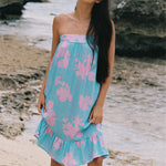 A woman stands on a sandy beach wearing a strapless light blue dress with a pink floral print. The background shows rocks and the ocean.