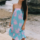 A woman stands on a sandy beach wearing a strapless light blue dress with a pink floral print. The background shows rocks and the ocean.