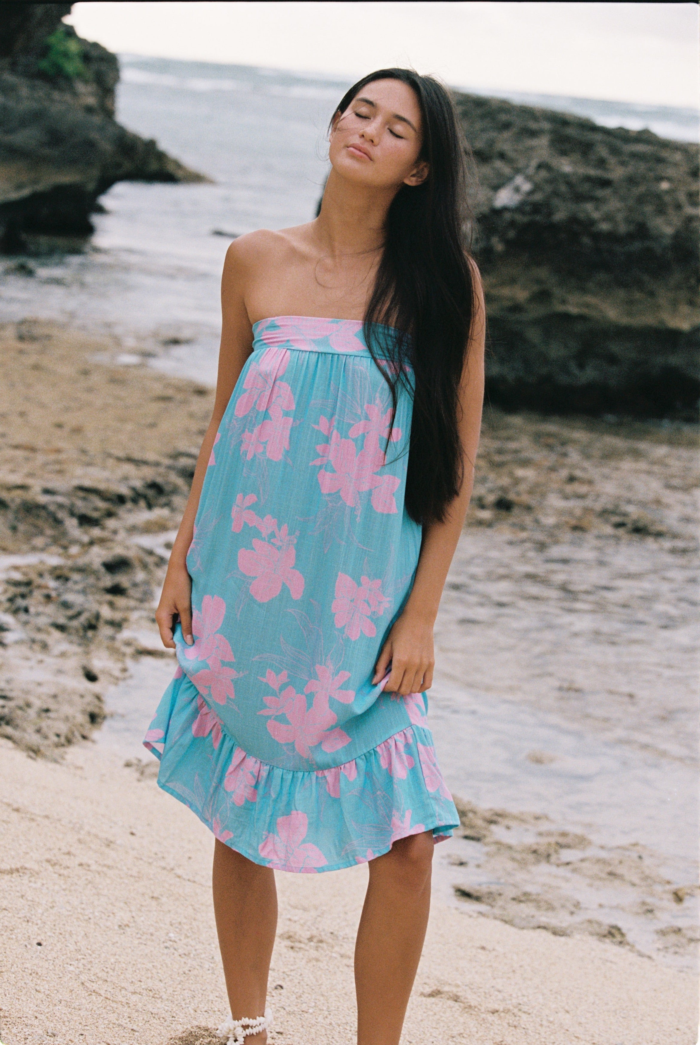 A woman stands on a sandy beach wearing a strapless light blue dress with a pink floral print. The background shows rocks and the ocean.