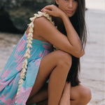 A woman crouching on a sandy beach wearing a light blue floral wrap dress with a white flower lei draped over her shoulder, with rocks and water in the background.