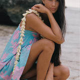 A woman crouching on a sandy beach wearing a light blue floral wrap dress with a white flower lei draped over her shoulder, with rocks and water in the background.