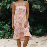 A woman stands barefoot on sandy ground in front of dense green foliage, wearing a strapless pink dress with a floral print and ruffled hem.