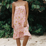 A woman stands barefoot on sandy ground in front of dense green foliage, wearing a strapless pink dress with a floral print and ruffled hem.