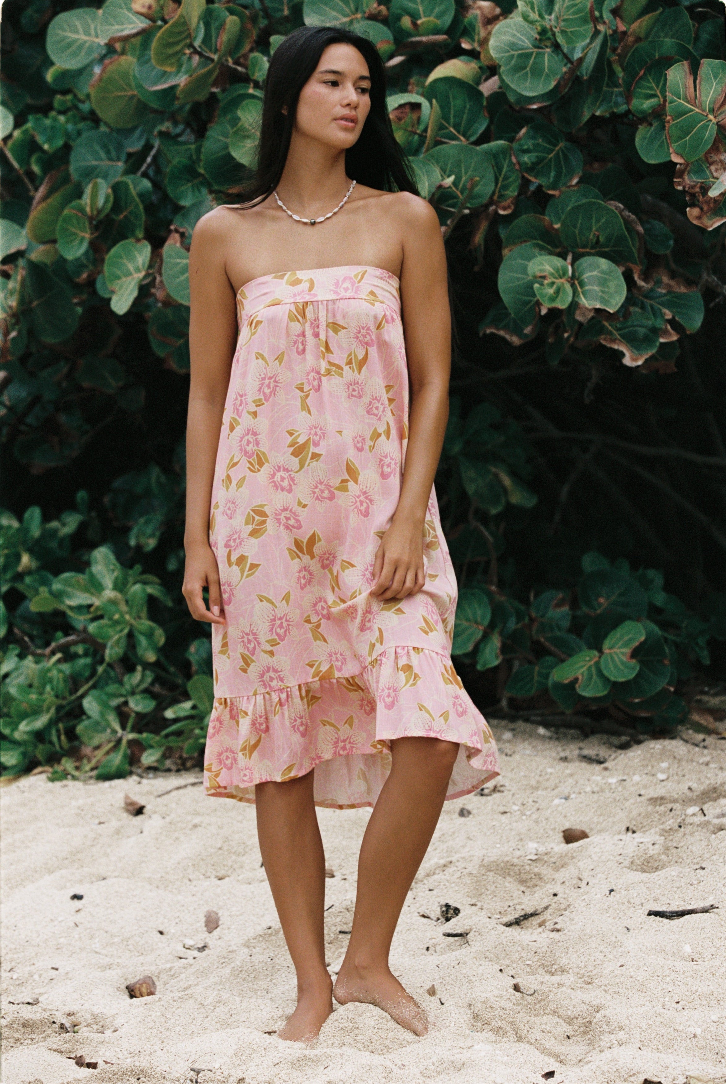 A woman stands barefoot on sandy ground in front of dense green foliage, wearing a strapless pink dress with a floral print and ruffled hem.