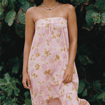 A woman wearing a strapless pink dress with a floral print, standing outdoors in front of a background of green tropical leaves.