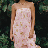 A woman wearing a strapless pink dress with a floral print, standing outdoors in front of a background of green tropical leaves.