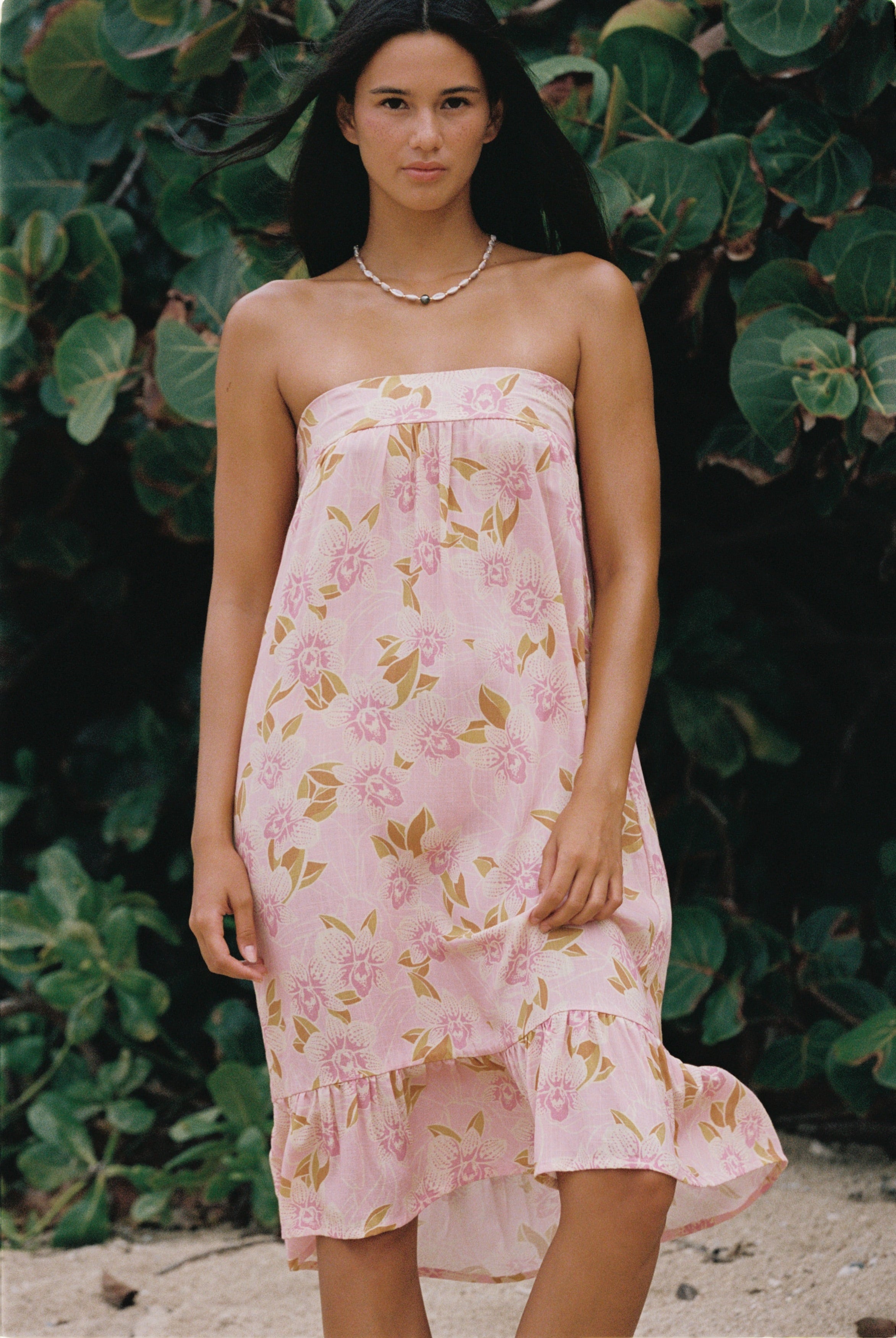 A woman wearing a strapless pink dress with a floral print, standing outdoors in front of a background of green tropical leaves.