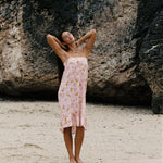 A woman stands on a sandy beach in front of a large rock formation, wearing a light pink floral strapless dress with a ruffled hem.