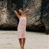 A woman stands on a sandy beach in front of a large rock formation, wearing a light pink floral strapless dress with a ruffled hem.