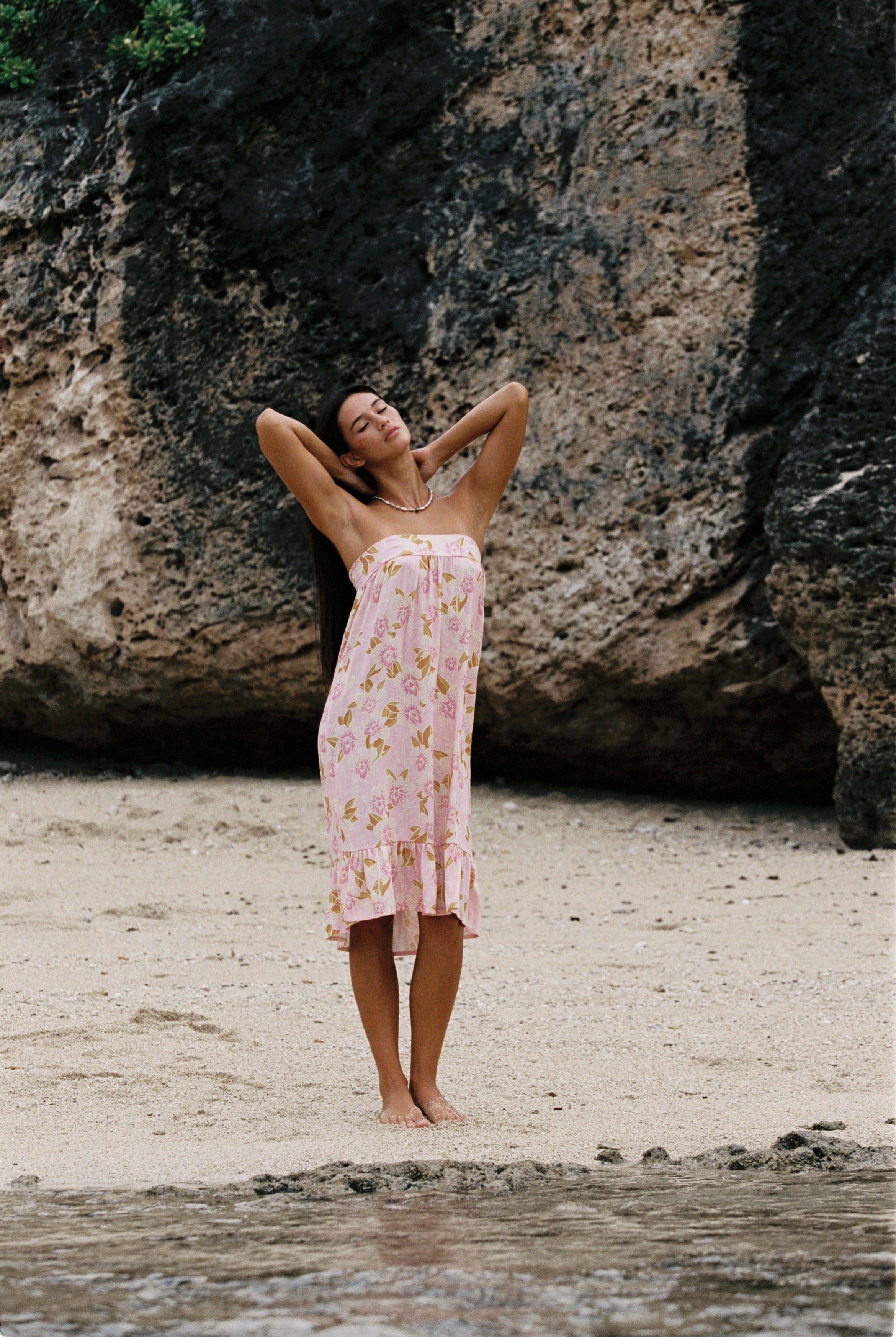 A woman stands on a sandy beach in front of a large rock formation, wearing a light pink floral strapless dress with a ruffled hem.