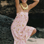 A woman posing on a beach or rocky shore wearing a light pink strapless dress with a yellow and green floral pattern, a yellow flower lei necklace, and pink sunglasses.