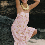 A woman posing on a beach or rocky shore wearing a light pink strapless dress with a yellow and green floral pattern, a yellow flower lei necklace, and pink sunglasses.