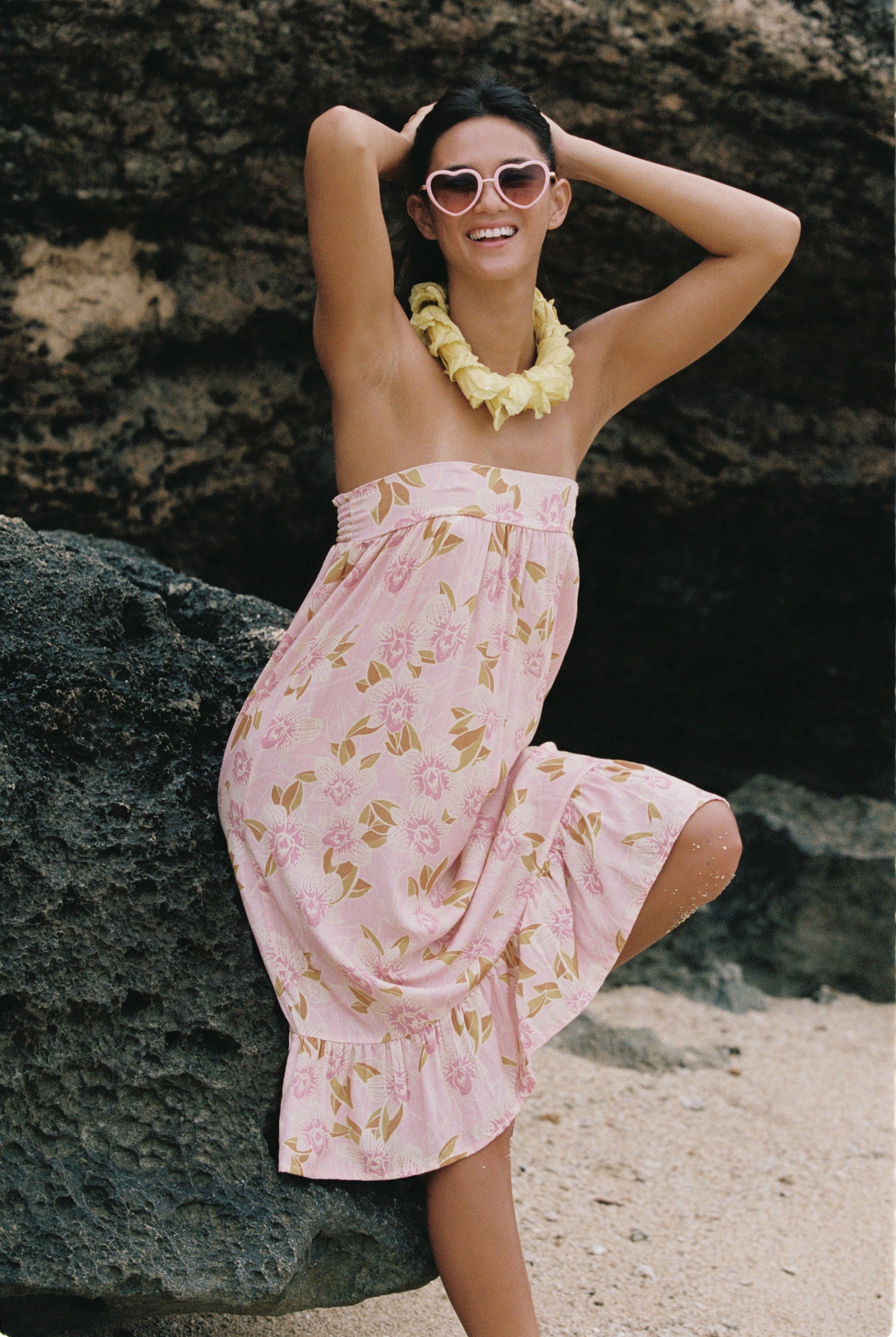 A woman posing on a beach or rocky shore wearing a light pink strapless dress with a yellow and green floral pattern, a yellow flower lei necklace, and pink sunglasses.