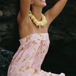 A woman wearing a light pink strapless dress with a gold and white floral pattern, a yellow flower lei necklace, and heart-shaped sunglasses, posing outdoors against a dark rocky background.