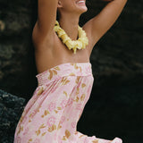 A woman wearing a light pink strapless dress with a gold and white floral pattern, a yellow flower lei necklace, and heart-shaped sunglasses, posing outdoors against a dark rocky background.
