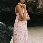 A woman wearing a light pink sleeveless dress with a yellow and green floral print, standing outdoors against a dark rock wall.