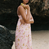A woman wearing a light pink sleeveless dress with a yellow and green floral print, standing outdoors against a dark rock wall.