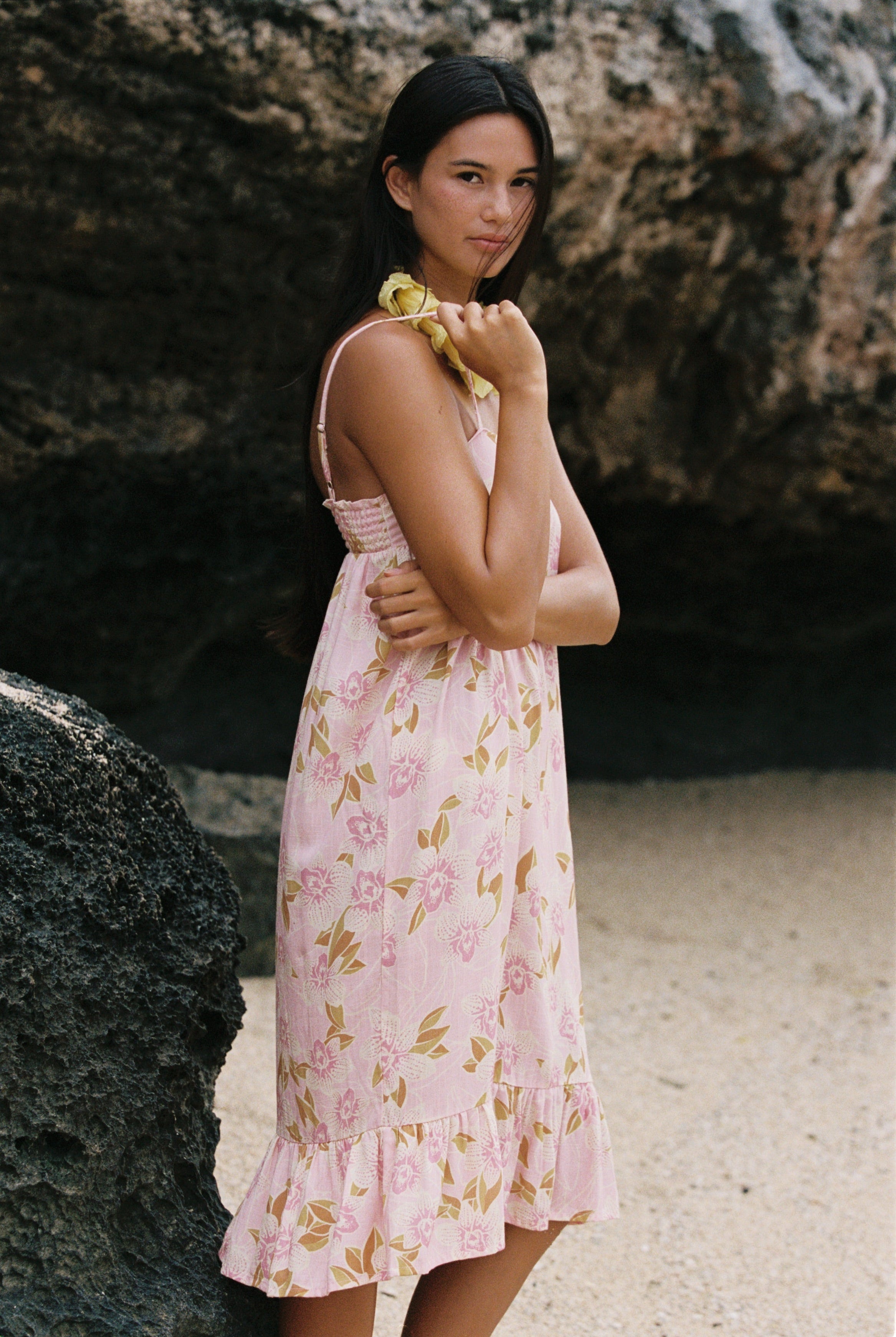 A woman wearing a light pink sleeveless dress with a yellow and green floral print, standing outdoors against a dark rock wall.