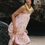 A woman wearing a light pink floral halter dress with a ruffled hem, standing on a sandy beach in front of a large rock formation. She is accessorized with a yellow flower lei necklace.
