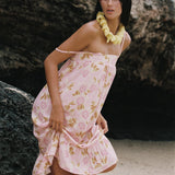 A woman wearing a light pink floral halter dress with a ruffled hem, standing on a sandy beach in front of a large rock formation. She is accessorized with a yellow flower lei necklace.