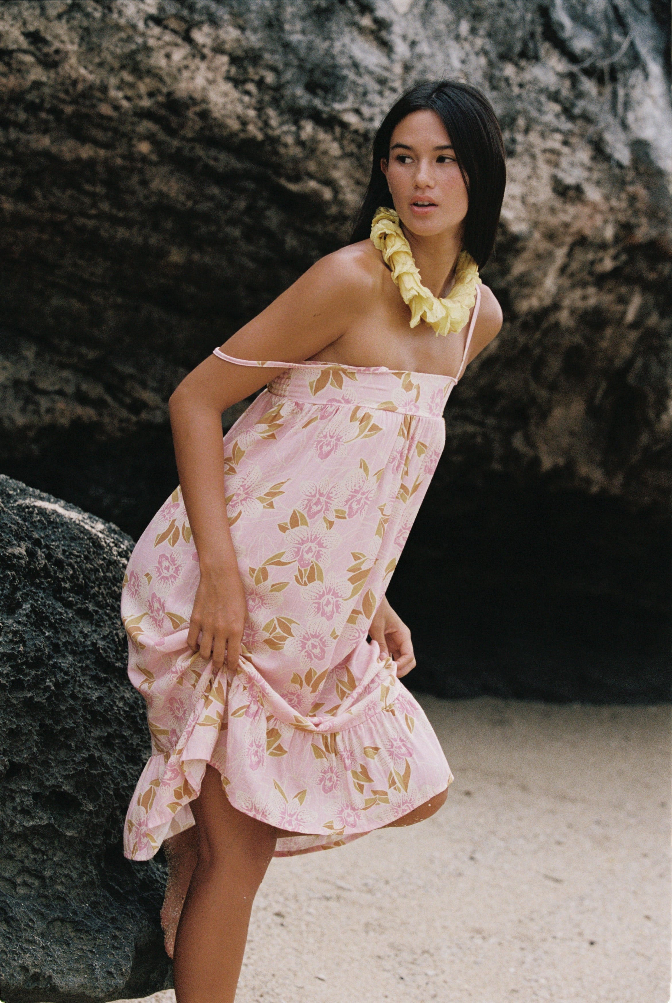 A woman wearing a light pink floral halter dress with a ruffled hem, standing on a sandy beach in front of a large rock formation. She is accessorized with a yellow flower lei necklace.