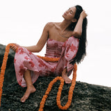 A woman sits barefoot on a dark rock against a bright white sky, wearing a pink floral strapless dress and draped orange flower garlands.