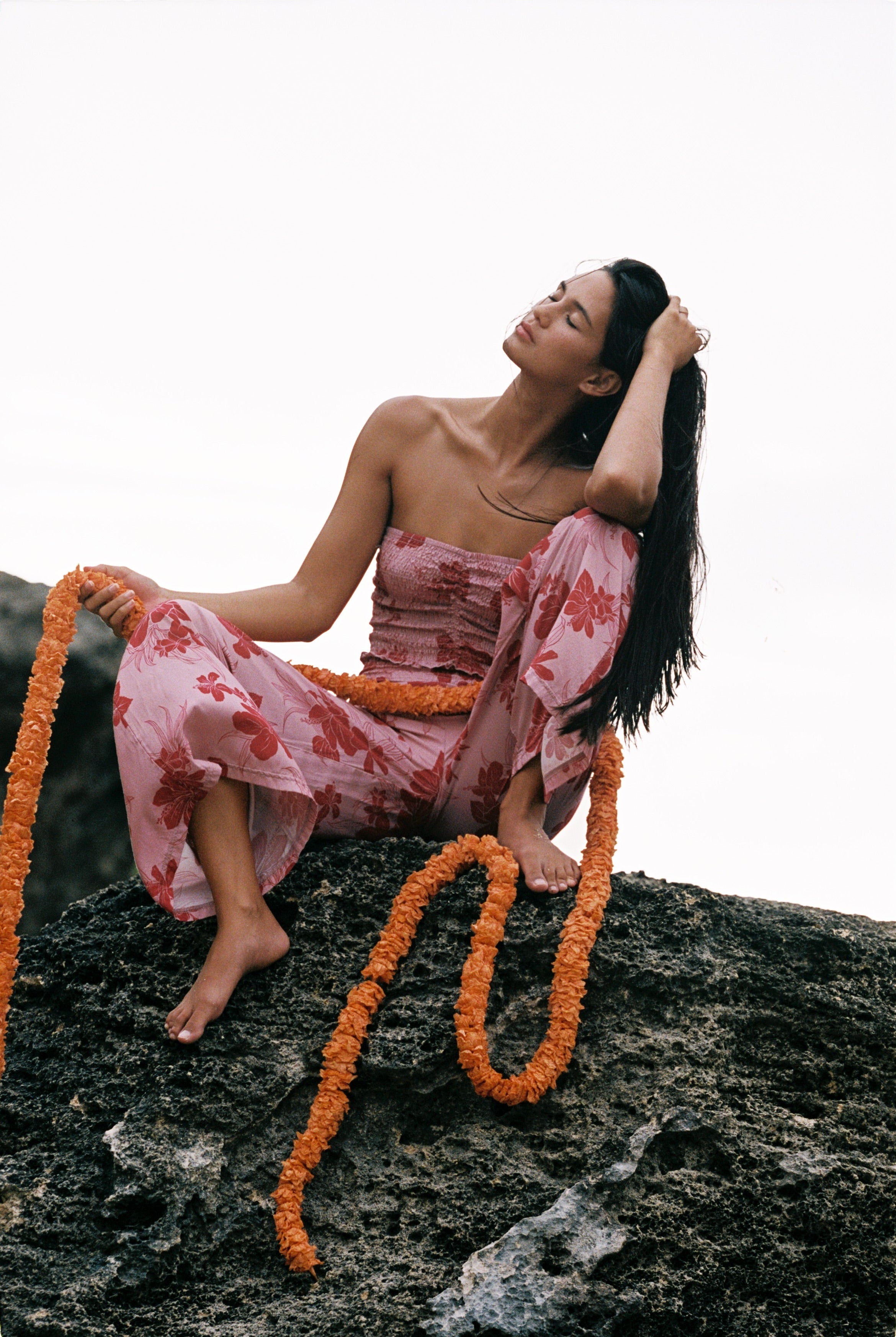 A woman sits barefoot on a dark rock against a bright white sky, wearing a pink floral strapless dress and draped orange flower garlands.
