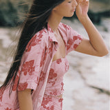 A woman wearing the solstice shirt by Guava & Co, a pink floral two-piece set with a matching open shirt, standing outdoors.