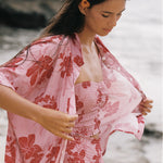 A woman on a beach wearing a light pink floral kimono-style cover-up over a matching pink smocked dress, with rocks and sand blurred in the background.