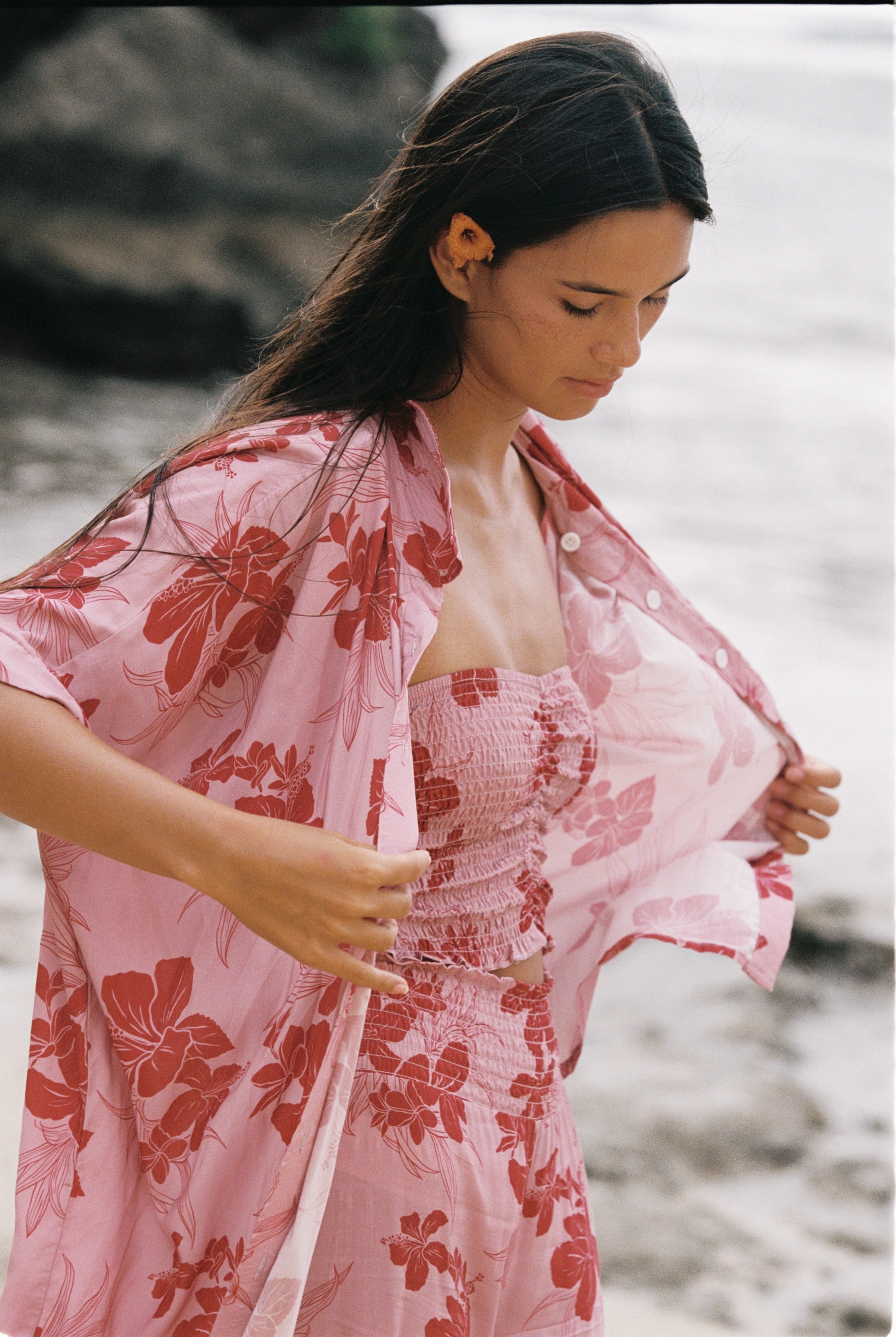 A woman on a beach wearing a light pink floral kimono-style cover-up over a matching pink smocked dress, with rocks and sand blurred in the background.