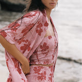A woman on a beach wearing a light pink two-piece set with a red floral print, including a smocked crop top and matching skirt, along with a matching open-front cardigan.