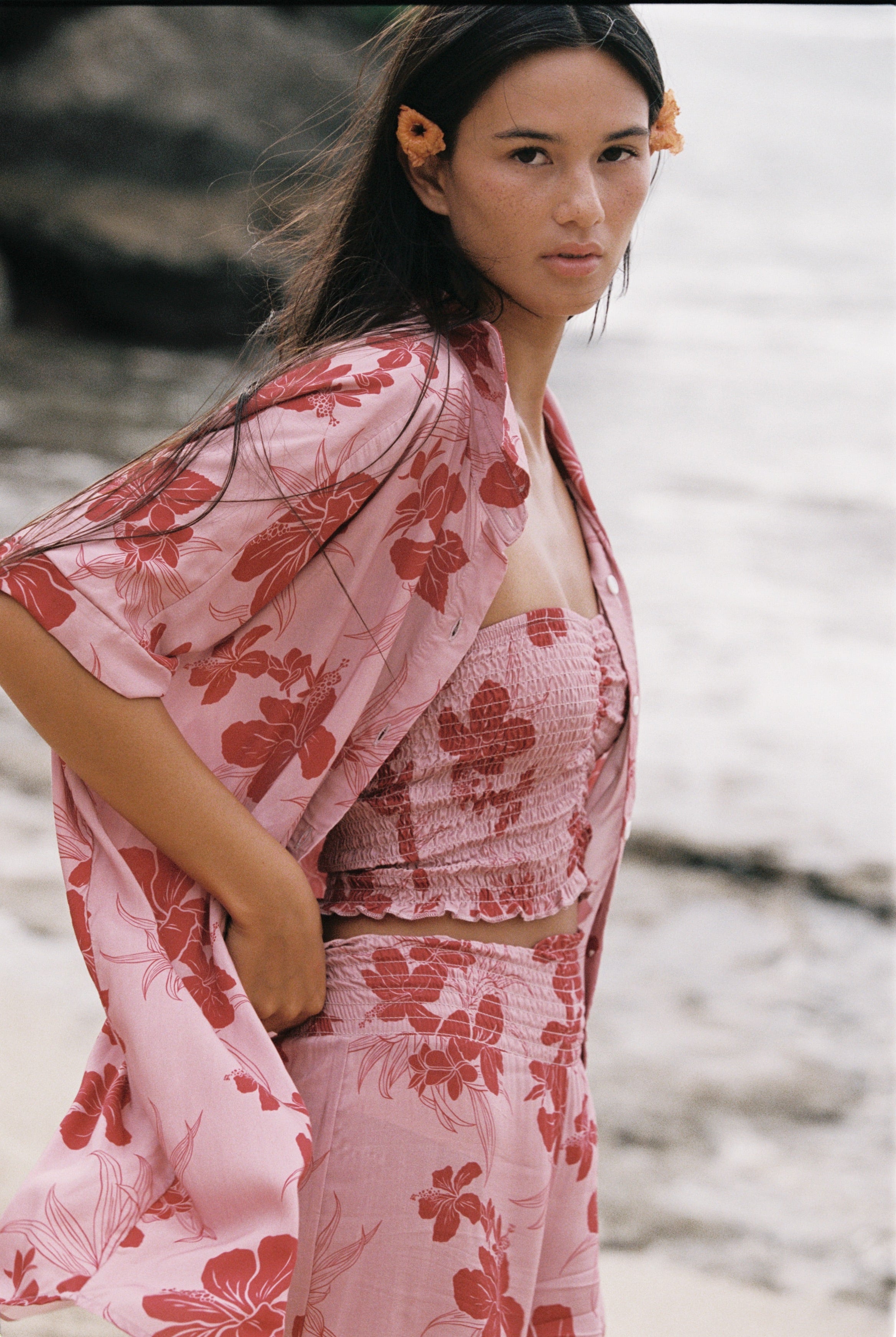 A woman on a beach wearing a light pink two-piece set with a red floral print, including a smocked crop top and matching skirt, along with a matching open-front cardigan.