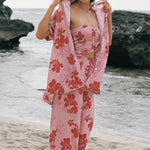 A woman stands on a sandy beach wearing a light pink floral two-piece set with a matching robe draped over her shoulders. The set features a strapless bandeau-style top and wide-leg pants with large red-orange hibiscus flowers on a pale pink background.