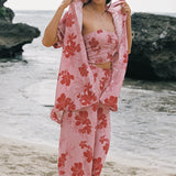 A woman stands on a sandy beach wearing a light pink floral two-piece set with a matching robe draped over her shoulders. The set features a strapless bandeau-style top and wide-leg pants with large red-orange hibiscus flowers on a pale pink background.