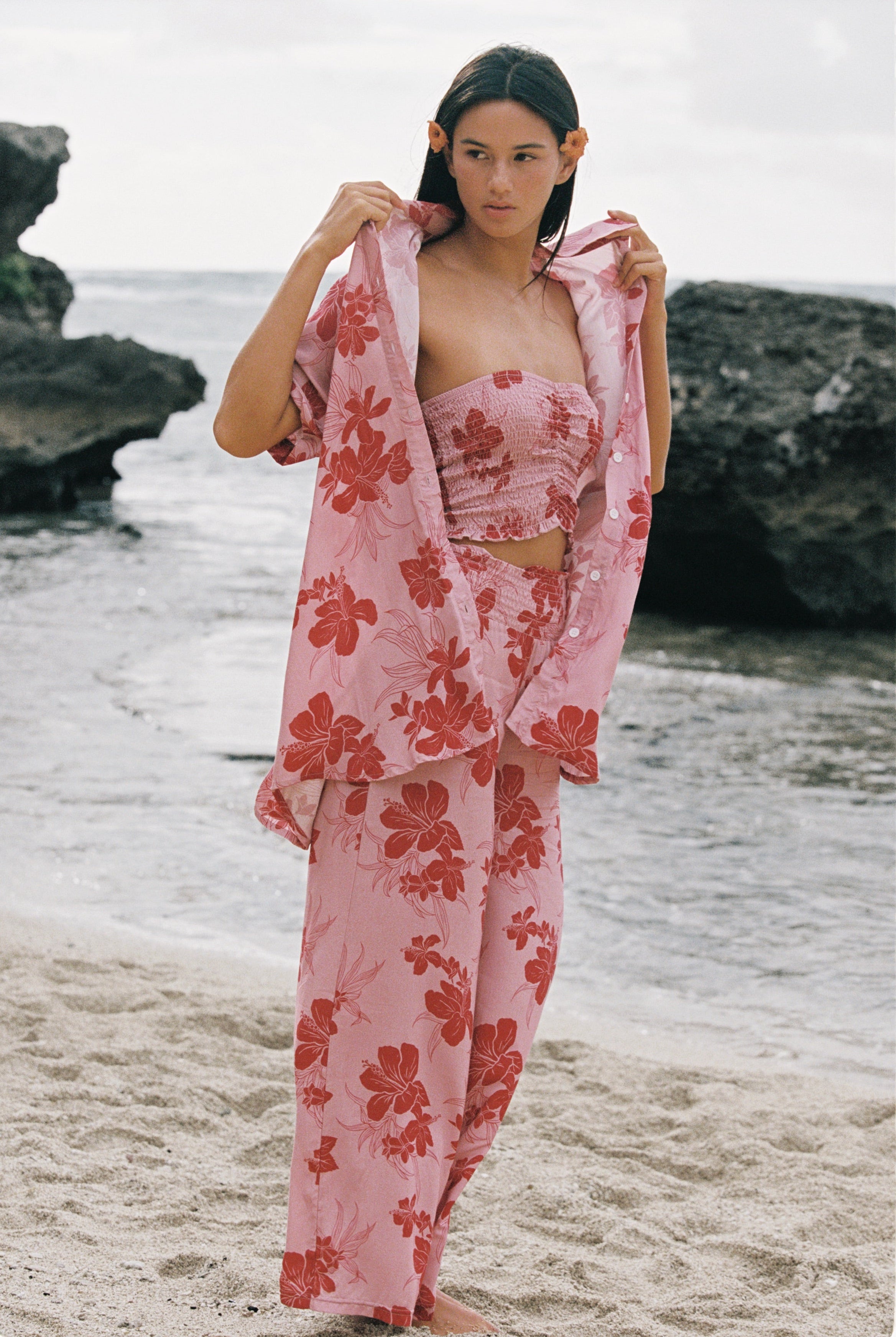 A woman stands on a sandy beach wearing a light pink floral two-piece set with a matching robe draped over her shoulders. The set features a strapless bandeau-style top and wide-leg pants with large red-orange hibiscus flowers on a pale pink background.