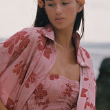 A woman standing outdoors near the water wearing a matching pink floral lounge set with a matching open shirt, including a strapless top and pants.