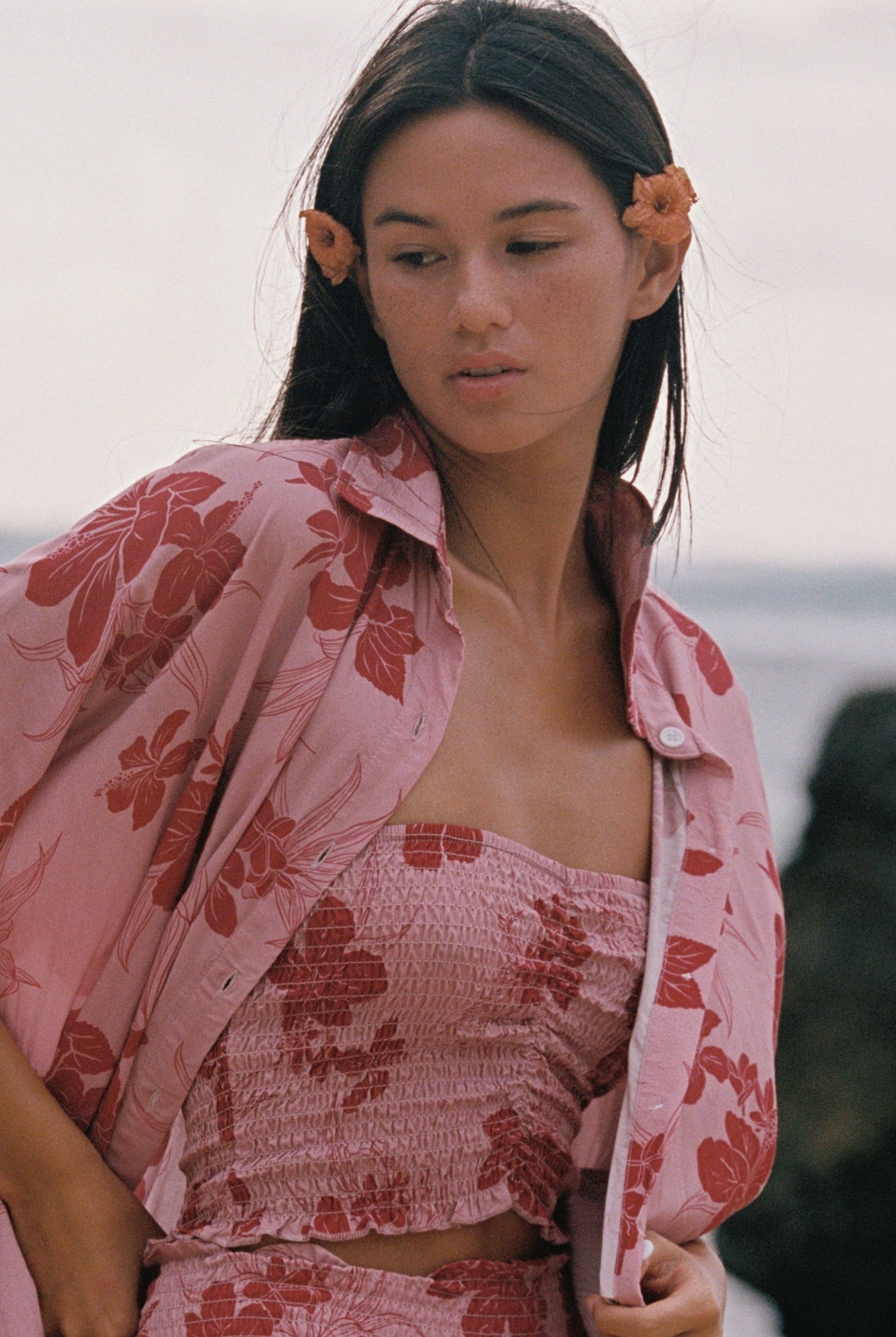 A woman standing outdoors near the water wearing a matching pink floral lounge set with a matching open shirt, including a strapless top and pants.
