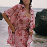 A woman standing outdoors near the water wearing a matching pink floral lounge set with a matching open shirt, including a strapless top and pants.