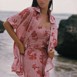 A woman standing outdoors near the water wearing a matching pink floral lounge set with a matching open shirt, including a strapless top and pants.