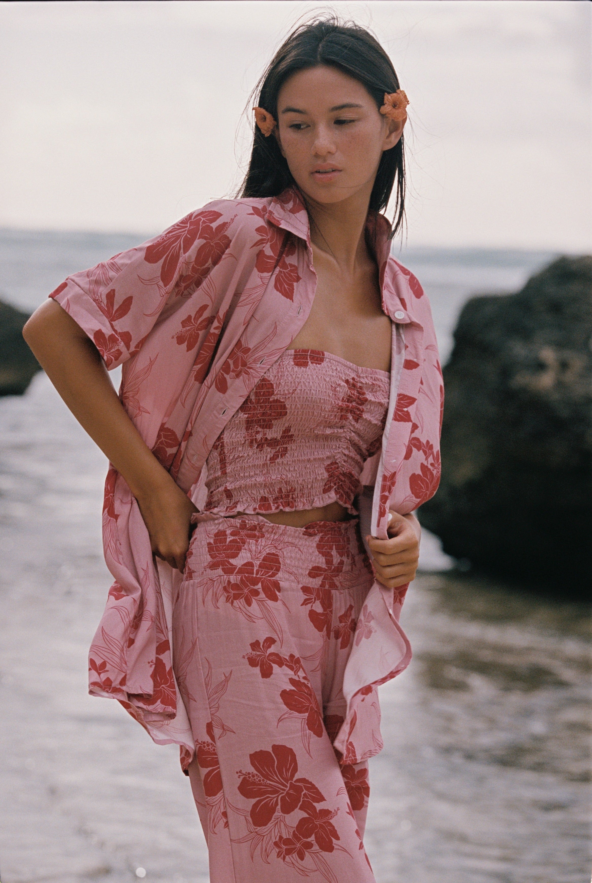 A woman standing outdoors near the water wearing a matching pink floral lounge set with a matching open shirt, including a strapless top and pants.