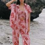 A woman stands on a beach wearing the solstice shirt by Guava & Co, a matching pink floral lounge set with a short-sleeve button-up shirt, cropped tank top, and wide-leg pants. Large dark rocks and shallow water are behind her.