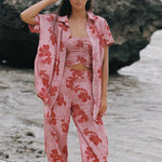 A woman stands on a beach wearing a matching pink floral lounge set with a short-sleeve button-up shirt, cropped tank top, and wide-leg pants. Large dark rocks and shallow water are behind her.