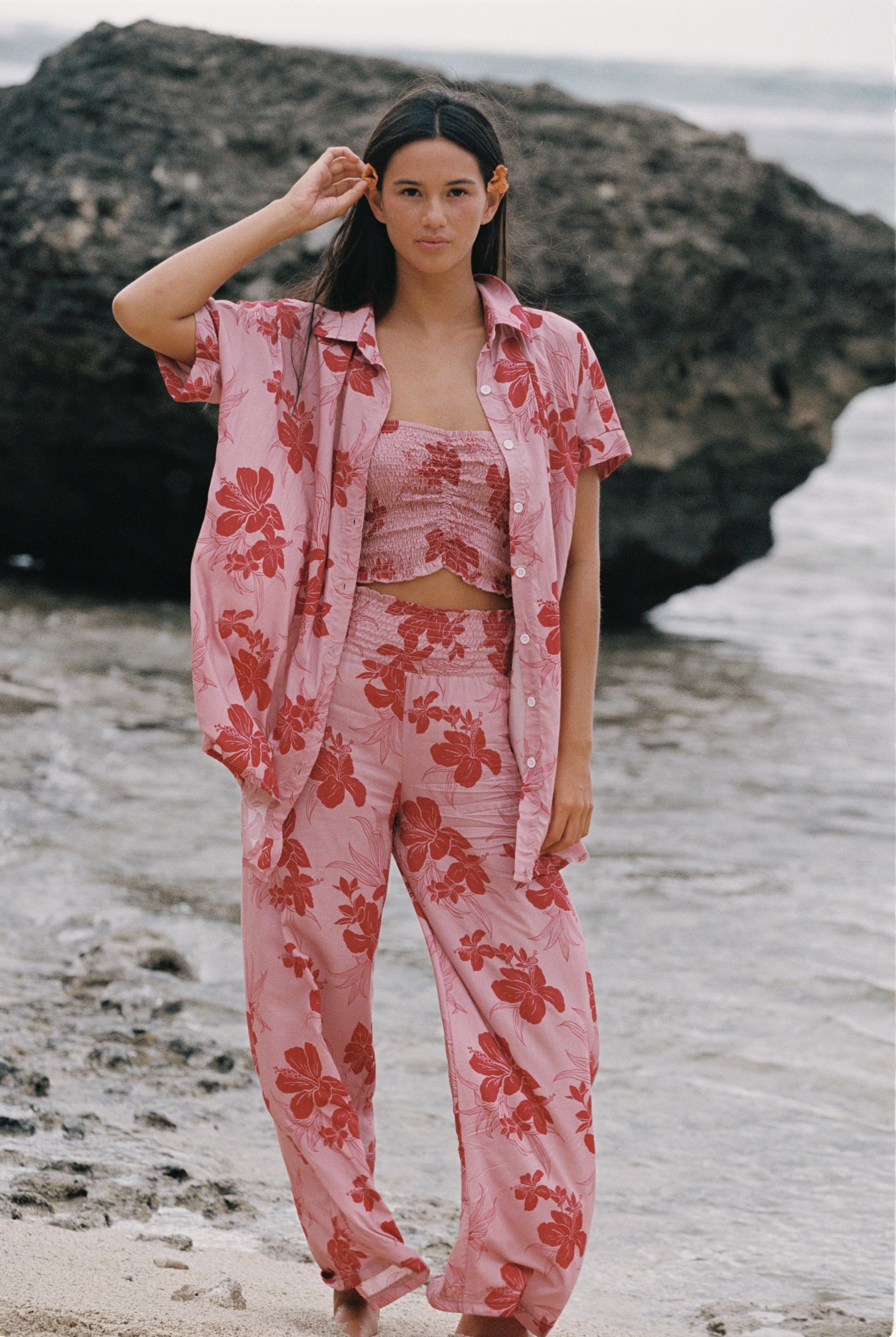 A woman stands on a beach wearing a matching pink floral lounge set with a short-sleeve button-up shirt, cropped tank top, and wide-leg pants. Large dark rocks and shallow water are behind her.